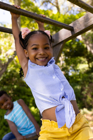Low angle view of african american cute smiling girl hanging on monkeys bars at playground....