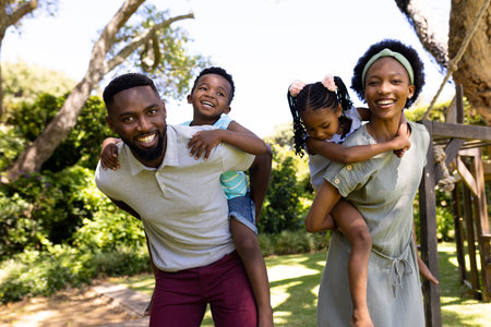 African american parents piggybacking playful son and daughter while standing in park, copy space. Unaltered, family, togetherness, childhood, enjoyment and weekend concept.の写真素材