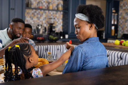 African american man and son looking at daughter feeding strawberry to mother at dining table. Unaltered, family, togetherness, childhood, breakfast, pancake, croissant, lifestyle and home concept.の写真素材