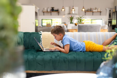 Side view of caucasian boy using laptop while lying on sofa in living room, copy space. Unaltered, lifestyle, childhood, relaxing, focus, technology and home concept.の写真素材
