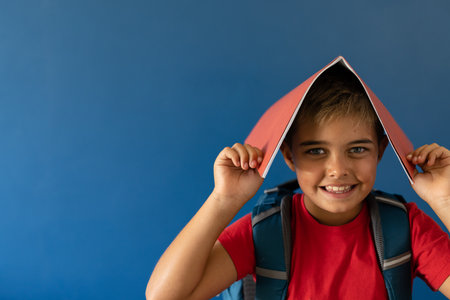 Portrait of playful caucasian boy with book on head standing over blue background, copy space. Unaltered, lifestyle, childhood, school boy, student and happy concept.の写真素材