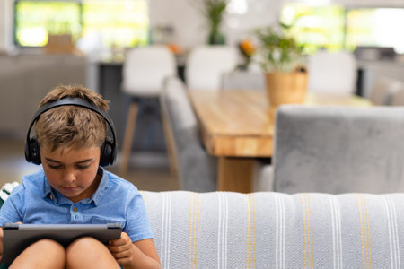 Caucasian boy wearing headphones and using digital tablet while relaxing on sofa in living room....