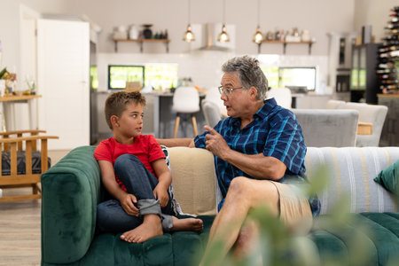 Caucasian senior man gesturing and talking with cute grandson while sitting on sofa in living room....