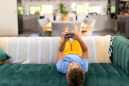 Caucasian boy using mobile phone while lying on sofa in living room, copy space. Unaltered, lifestyle, childhood, relaxing, technology and home concept.の写真素材
