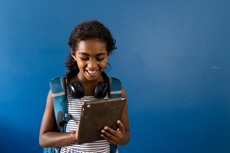 Happy biracial schoolgirl with headphones using tablet, smiling on blue background with copy space. Childhood, education, school and technology.の写真素材