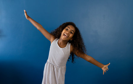 Portrait of happy biracial girl with arms outstretched laughing, on blue background with copy space....
