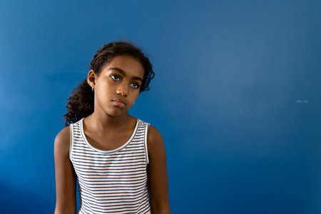 Portrait of sad biracial girl wearing striped dress on blue background with copy space. Studio shot, childhood, emotion and sadness.の写真素材