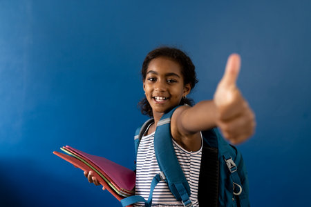 Portrait of happy biracial schoolgirl making thumbs up sign holding notebooks with copy space. Studio shot, childhood, education, learning and positivity.の写真素材