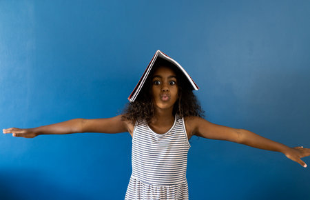 Portrait of biracial schoolgirl with arms outstretched and notebook on head, on blue background. Studio shot, childhood, education, learning and fun.の写真素材