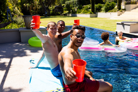 Portrait of happy diverse group of friends having pool party, holding plastic cups in garden....