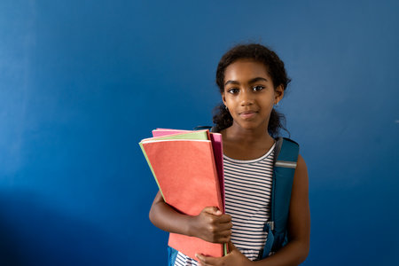 Portrait of happy biracial schoolgirl holding notebooks on blue background with copy space. Studio shot, childhood, education, learning and joy.の写真素材