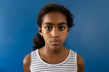 Portrait of sad biracial girl wearing striped dress on blue background with copy space. Studio shot, childhood, emotion and sadness.の写真素材
