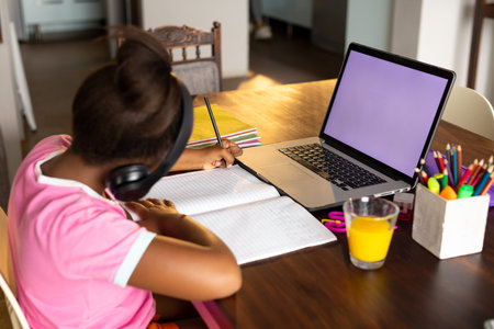 African american girl doing schoolwork using headphones and laptop at home, copy space on screen. Childhood, technology, education, online learning and domestic life.の写真素材