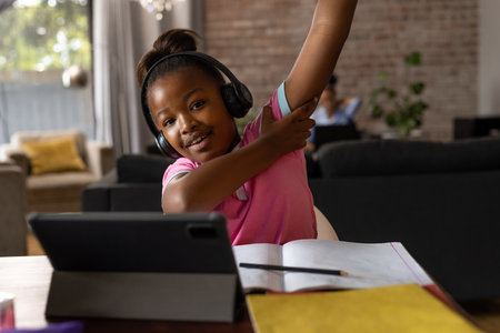 Happy african american girl wearing headphones raising hand during online lesson at home. Childhood,...