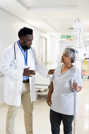 African american male doctor talking to diverse senior female patient with drip standing in corridor. Hospital, medical and healthcare services.の写真素材