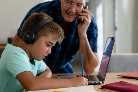Caucasian grandfather talking over phone and looking at grandson writing homework by laptop....