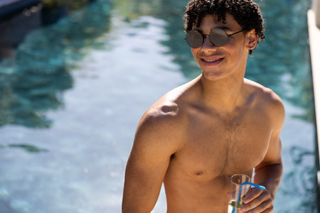 Happy shirtless biracial fit man in sunglasses drinking cocktail standing in swimming pool in sun. Summer, free time, relaxation and vacations.の写真素材
