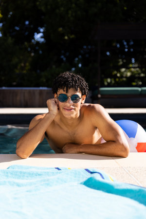 Portrait of happy biracial fit man in sunglasses standing in swimming pool in the sun, copy space. Summer, free time, relaxation and vacations.の写真素材
