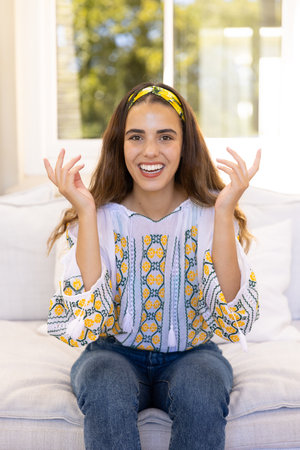 Biracial cheerful young woman gesturing and looking at camera while sitting on sofa in living room....