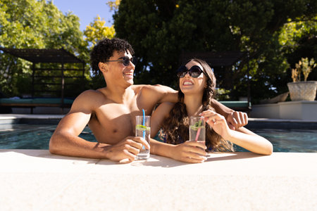 Happy diverse fit couple standing in swimming pool laughing in the sun with cocktails. Summer, free...