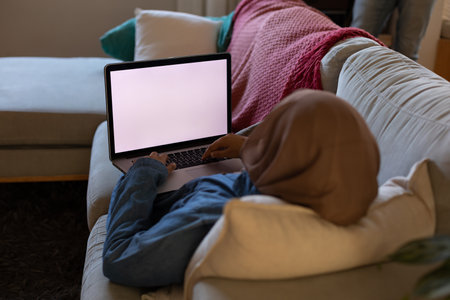 Biracial woman wearing hijab, laying on sofa in living room and using laptop with copy space. Spending quality time at home alone.の写真素材