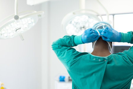 Rear view of african american surgeon in gown and gloves tying on mask in theatre, copy space. Hospital, medical and healthcare services.の写真素材