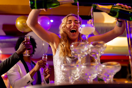 Happy caucasian woman pouring a champagne fountain into glasses with diverse friends at a nightclub....