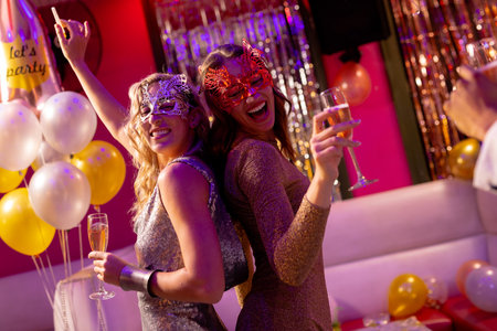 Two happy caucasian female friends in masks dancing and drinking champagne at a party in a nightclub. Fun, going out, celebrating and party concept.の写真素材