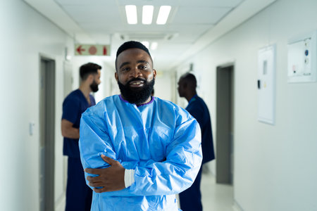 Portrait of smiling african american male healthcare worker in surgical gown in busy corridor. Hospital, medical and healthcare services.の写真素材