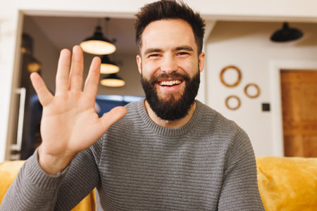 Portrait of happy biracial bearded young man waving hand while sitting on sofa in living room. Happy, gesturing, handsome, unaltered, lifestyle and home concept.の写真素材