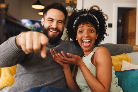 Portrait of cheerful biracial couple laughing and gesturing while sitting on sofa in living room....