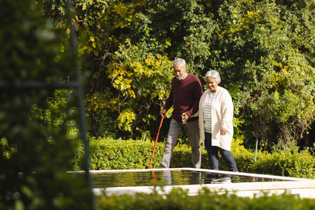 Happy senior biracial couple holding hands and walking with cane in garden at home. Senior lifestyle, retirement, nature and domestic life, unaltered.の写真素材