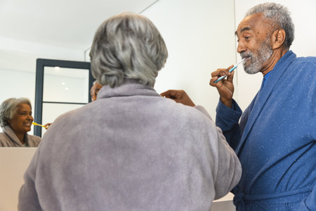 Happy senior biracial couple wearing bathrobes and brushing teeth in bathroom at home. Senior lifestyle, retirement, selfcare and domestic life, unaltered.の写真素材