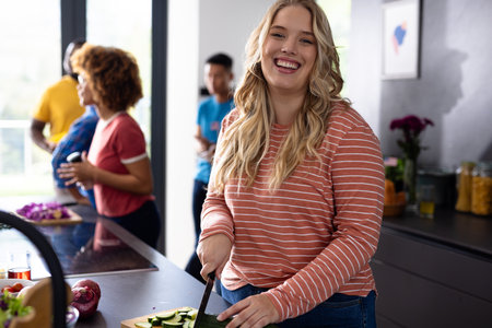 Portrait of happy caucasian woman cutting vegetables with diverse friends in kitchen. Domestic life, friendship, togetherness, food and lifestyle, unaltered.の写真素材