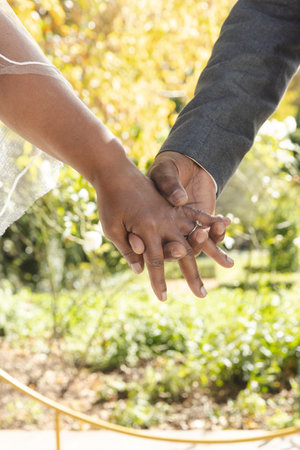 Midsection of senior biracial bride and groom holding hands at sunny outdoor wedding ceremony. Marriage, romance, love, tradition, ceremony, summer, retirement and senior lifestyle, unaltered.の写真素材