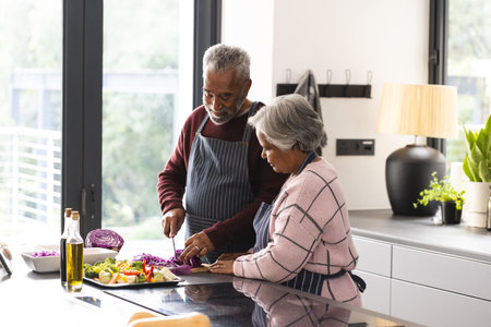 Happy senior biracial couple wearing aprons preparing vegetables in kitchen at home. Senior lifestyle, retirement, food, cooking and domestic life, unaltered.の写真素材