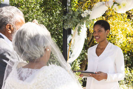 Biracial female marriage officiant using tablet with senior couple during wedding in sunny garden. Love, ceremony, marriage, summer and celebration, unaltered.の写真素材