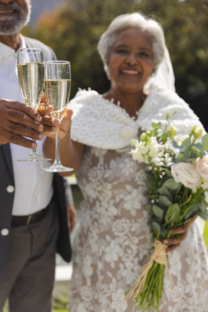 Happy senior biracial bride and groom drinking champagne toast in garden at sunny wedding ceremony. Marriage, romance, retirement and senior lifestyle, unaltered.の写真素材