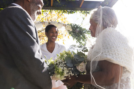 Happy biracial female marriage officiant with senior couple during wedding ceremony in sunny garden. Love, ceremony, marriage, summer and celebration, unaltered.の写真素材