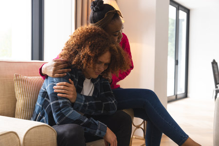 Sad diverse couple sitting in living room at home, embracing and comforting. Depression, mental health, relationship, support, togetherness, lifestyle and domestic life, unaltered.の写真素材