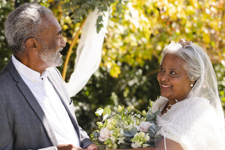 Happy senior biracial bride and groom during wedding ceremony in sunny garden. Marriage, love, romance, ceremony, summer, tradition and senior lifestyle, unaltered.の写真素材