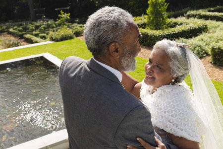 Happy senior biracial bride and groom dancing in garden at sunny wedding ceremony. Marriage, romance, love, tradition, ceremony, summer, retirement and senior lifestyle, unaltered.の写真素材