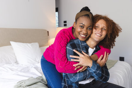 Portrait of happy diverse couple sitting on bed embracing in sunny bedroom at home, copy space. Relationship, togetherness, love, lifestyle, wellbeing and domestic life, unaltered.の写真素材