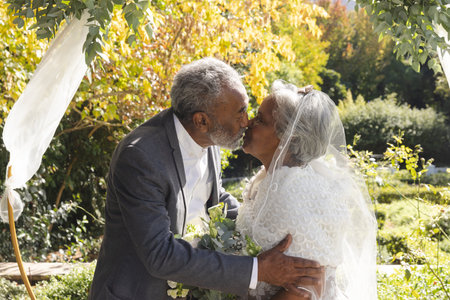 Happy senior biracial bride and groom kissing at sunny outdoor wedding ceremony.の写真素材