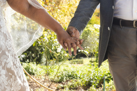 Midsection of senior biracial bride and groom holding hands at sunny outdoor wedding ceremony.の写真素材