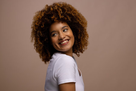 Happy biracial woman with dark curly hair smiling on brown background with copy space. Femininity,...