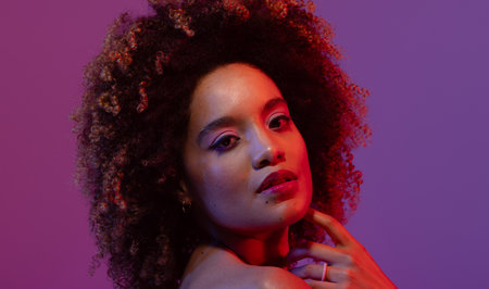 Biracial woman with curly hair touching chin on purple background. Femininity, face, facial...