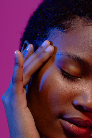 African american woman with eye closed touching head in blue light on purple background. Femininity, face, facial expressions, body, skin, makeup, fashion and beauty, unaltered.の写真素材
