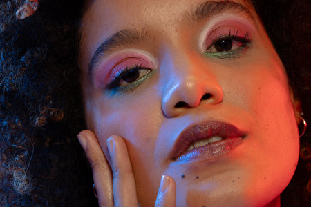 Close up of biracial woman with curly hair wearing eyeshadow and lipstick touching face. Femininity, face, facial expressions, body, skin, makeup, fashion and beauty, unaltered.の写真素材