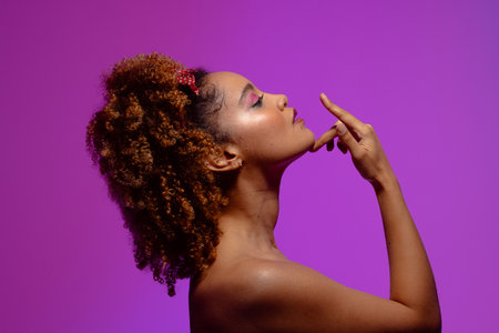 Biracial woman with pink eye shadow and lipstick touching chin, purple background. Femininity, face, facial expressions, body, skin, makeup, fashion and beauty, unaltered.の写真素材